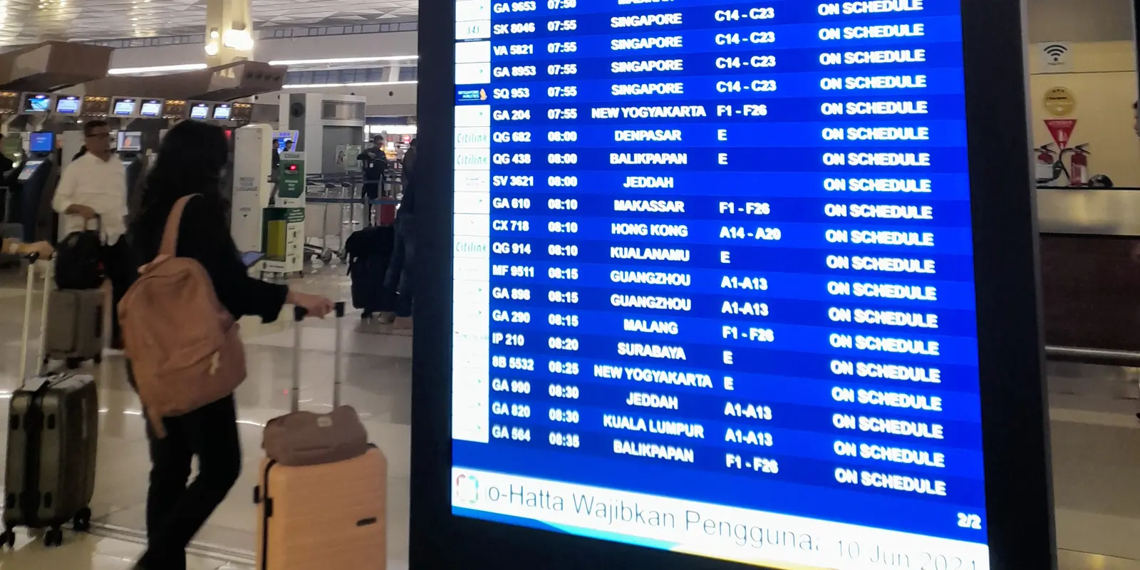 A tall flight information screen listing morning departures with the status "On schedule", beside travellers pulling suitcases through the check-in hall of Terminal 3 at Soekarno-Hatta.
