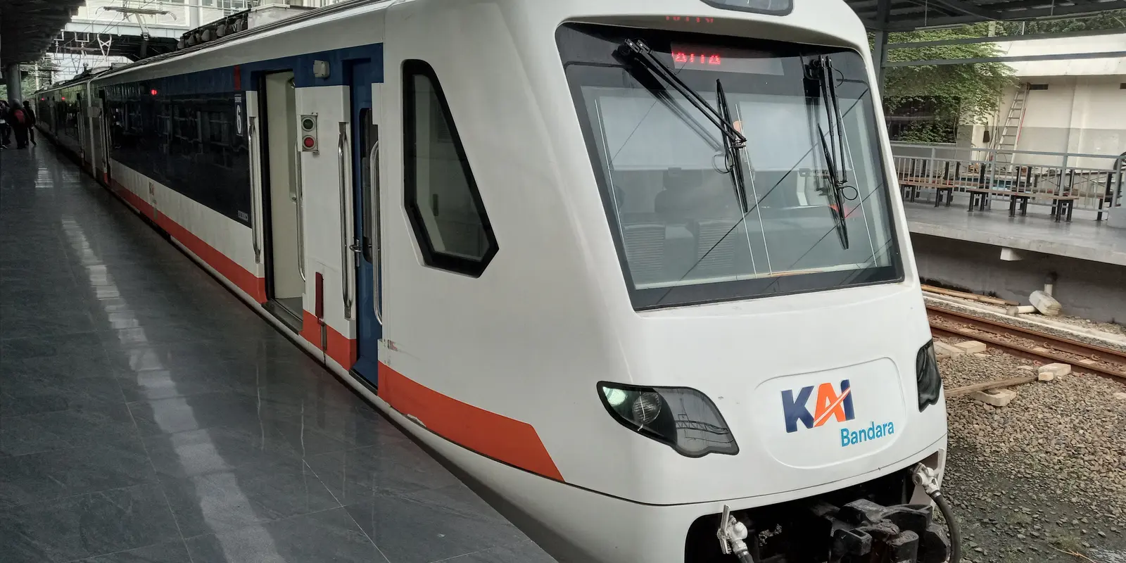 Rangkaian kereta bandara Soekarno-Hatta berwarna putih dengan garis merah dan biru serta logo KAI Bandara di bagian depan, berhenti di peron beratap Stasiun Batu Ceper. Beberapa penumpang menunggu di ujung peron; jalur kedua dan peron seberang terlihat di sebelah kanan.