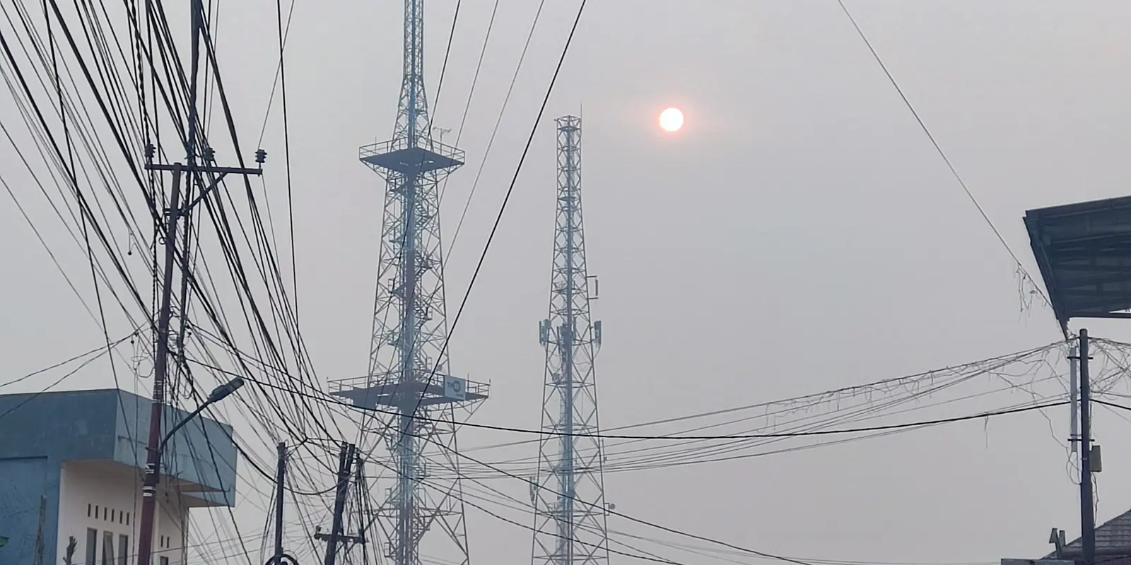 Two steel lattice transmission towers and a tangle of overhead power lines against a flat grey sky, with the sun showing as a small dim orange disc through thick smoke haze in Jambi City.