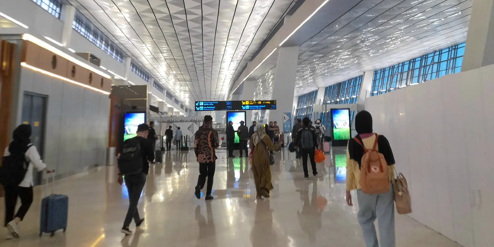 Passengers walking along a wide, brightly lit departure concourse at Soekarno-Hatta International Airport, with a faceted white ceiling, flight information screens and a polished stone floor.